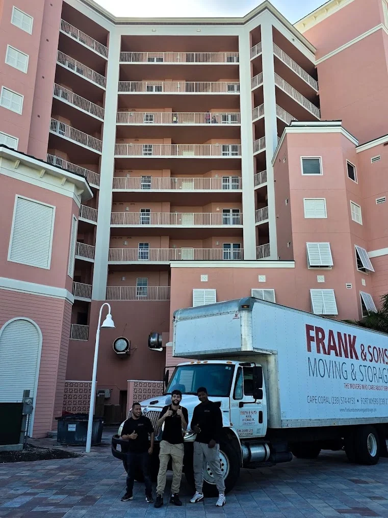 Three Frank and Sons crew members beside a moving truck and a pink condominium building